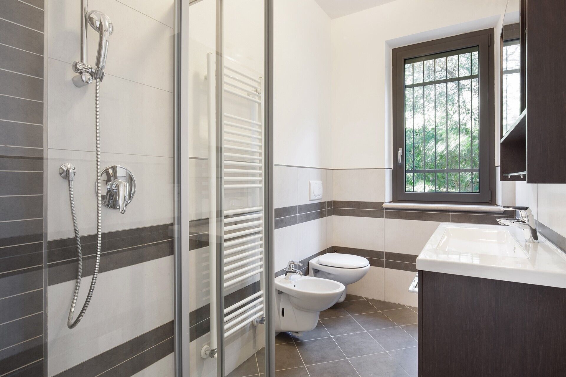 Une salle de bain moderne avec une douche à l’italienne en verre, des toilettes, un bidet et une fenêtre donnant sur un espace verdoyant.