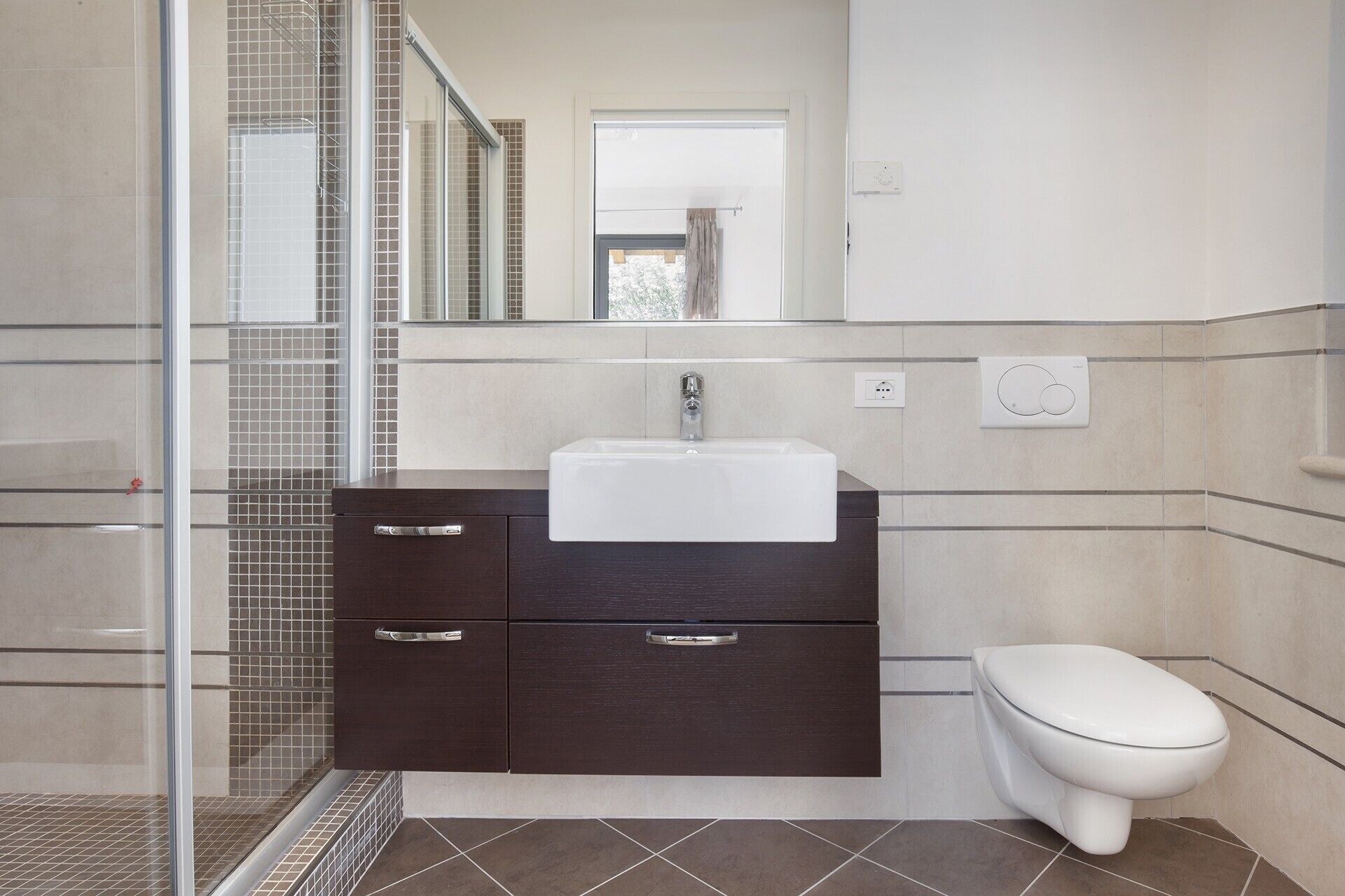 A bathroom with a glass shower enclosure, a white sink, dark cabinets, a toilet, and a mirror above the sink.