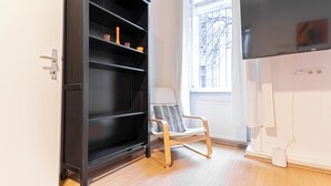Una stanza con un'unità di scaffalature nere, una sedia bianca, una TV a schermo piatto e una finestra che si affaccia su degli alberi.