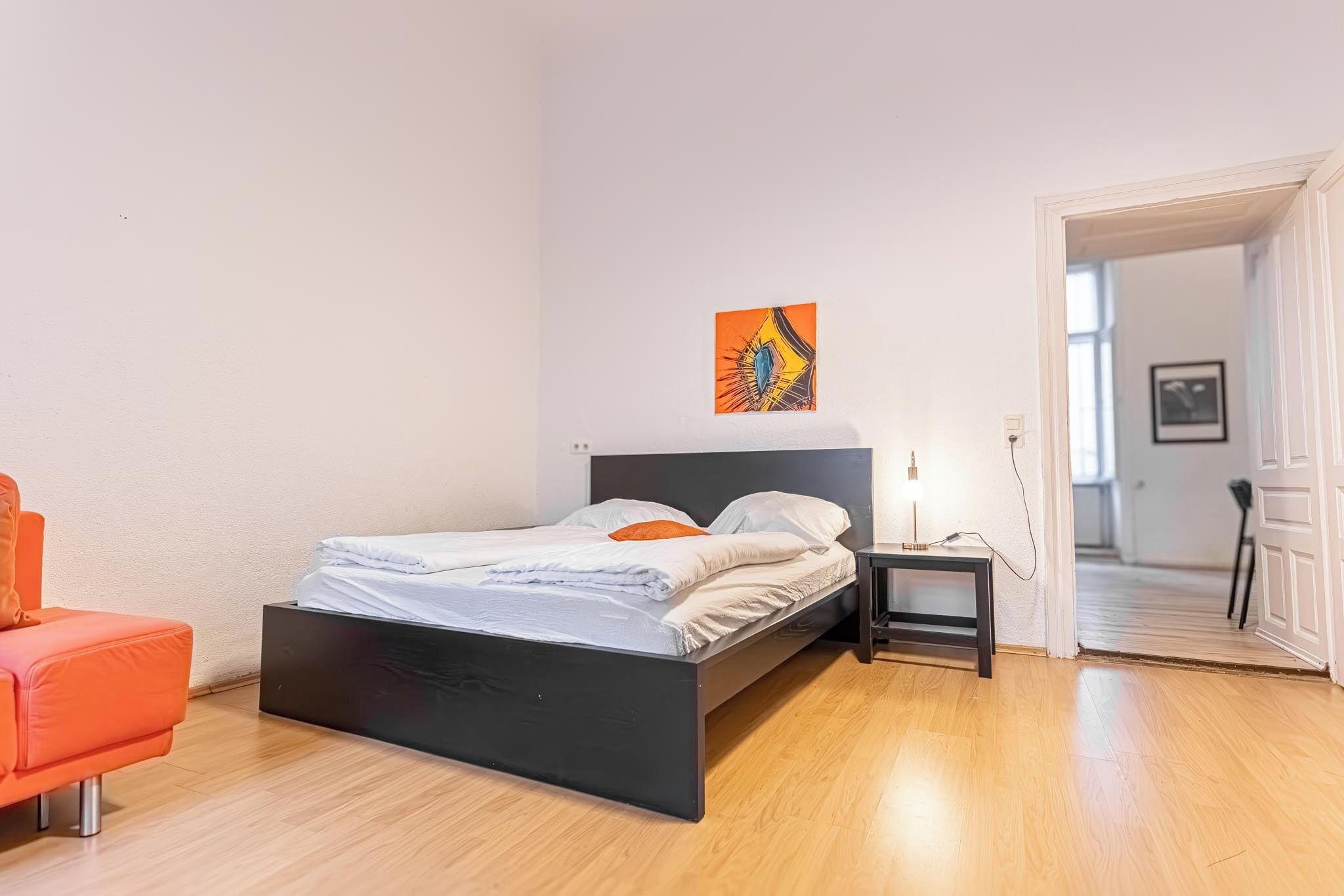 Ein modernes Schlafzimmer mit schwarzem Bettgestell, weißer Bettwäsche und einem orangefarbenen Sessel. Ein kleiner Nachttisch mit Lampe ist zu sehen.