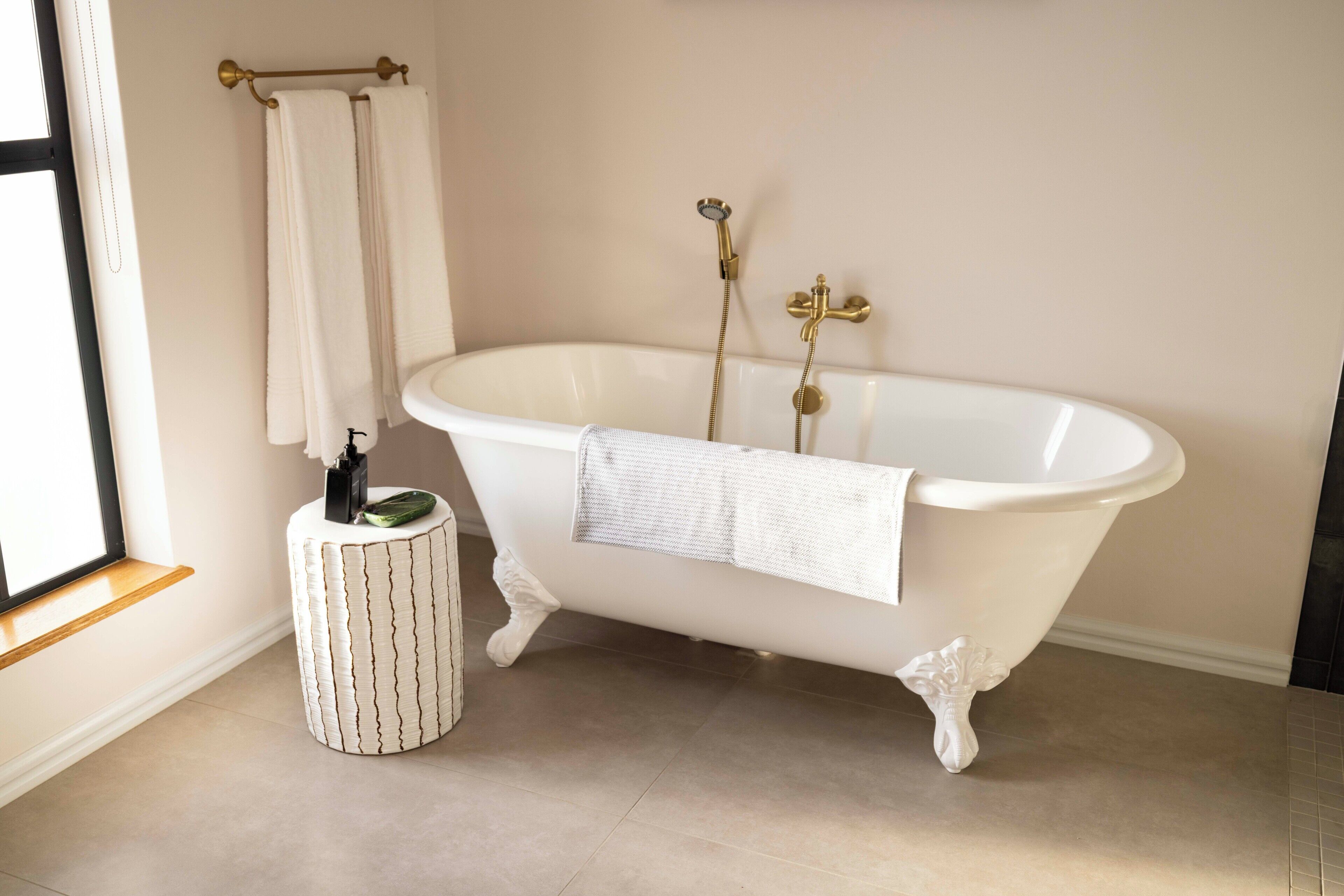 A bathroom with a freestanding bathtub, a towel rack with two towels, a small white stool, and a window with a wooden ledge.
