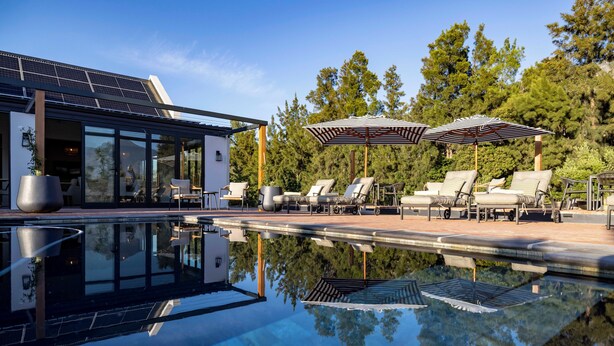 A modern outdoor pool area with lounge chairs and umbrellas, surrounded by trees and a building with solar panels. - Diggers Country House (Tulbagh)