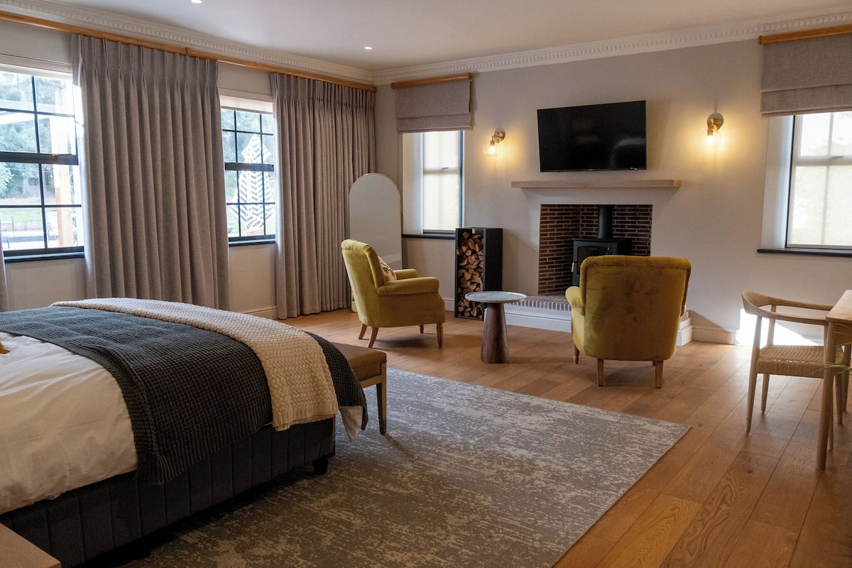 A hotel room with a large bed, two armchairs, a small table, a fireplace, and a TV mounted on the wall.