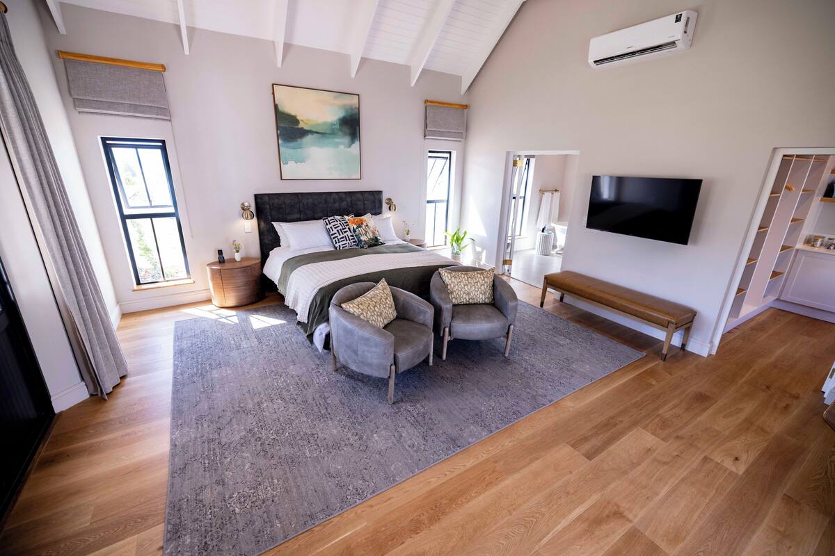A modern bedroom with a large bed, two armchairs, a bench, a flat-screen TV, and a large window.