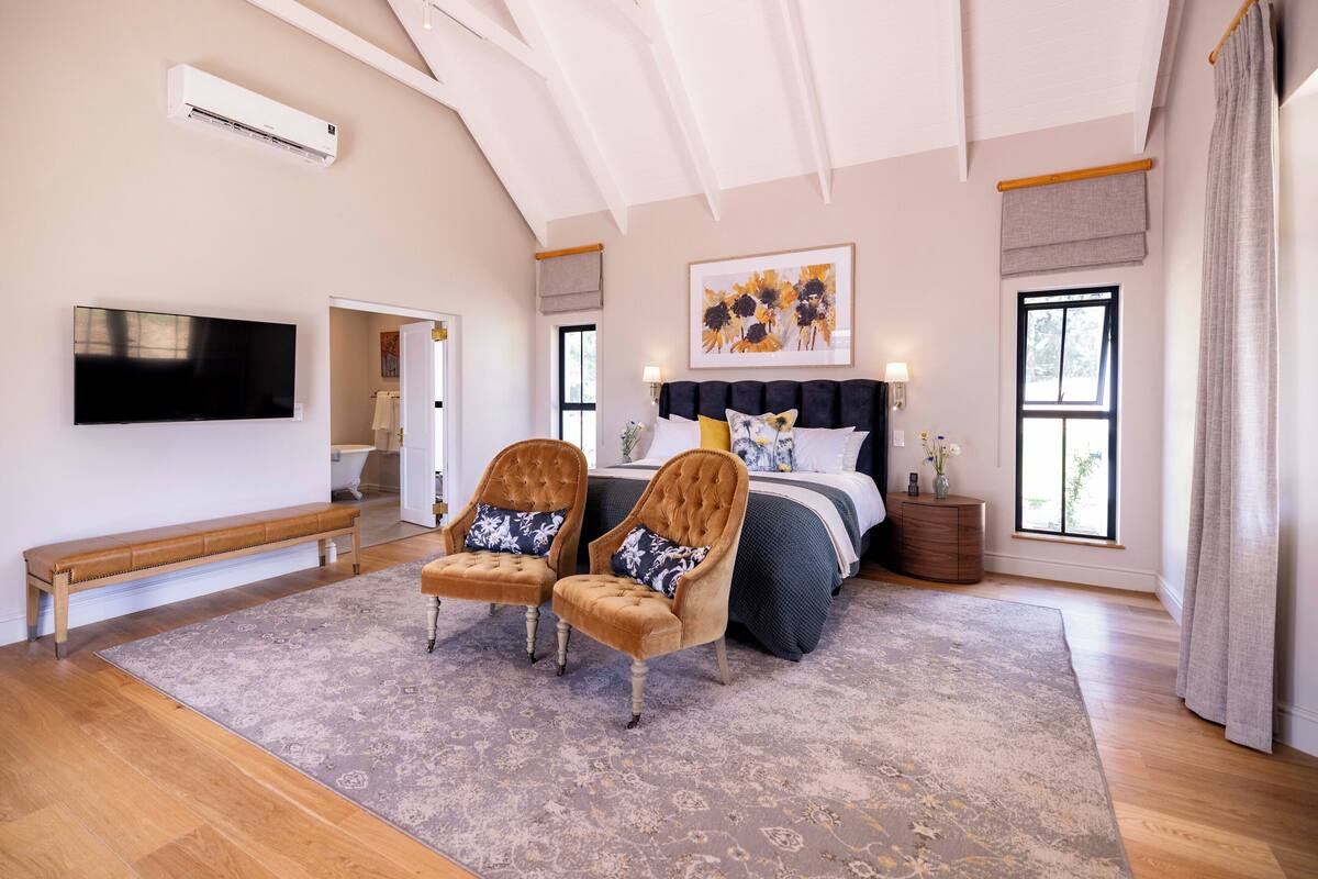 A bedroom with a large bed, two armchairs, a bench, a flat-screen TV, and a ceiling fan.