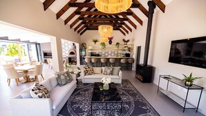 A modern living room with a sofa, coffee table, and patterned rug. There is a kitchen area with bar stools and a TV mounted on the wall. - Diggers Country House (Tulbagh)