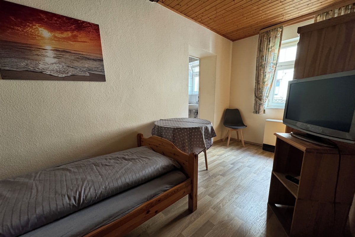Ett litet, ombonat rum med en säng i trä, ett runt bord, en tv på ett träställ och en tavla med en solnedgång på väggen.