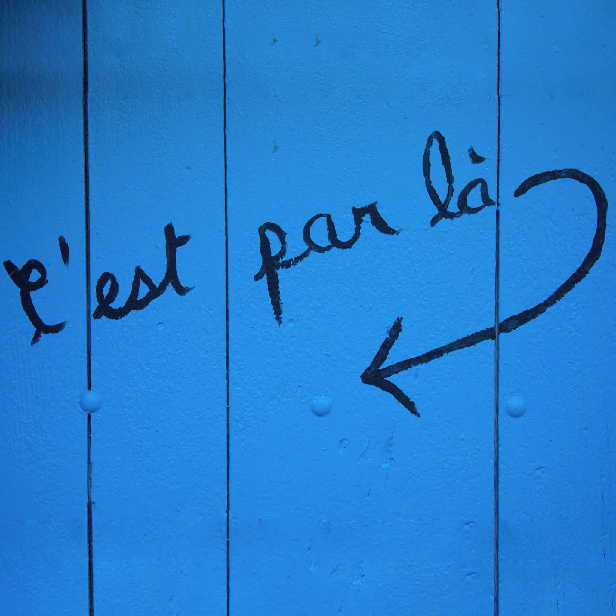 Pared azul con el texto "l'est par là" y una flecha apuntando a la izquierda.