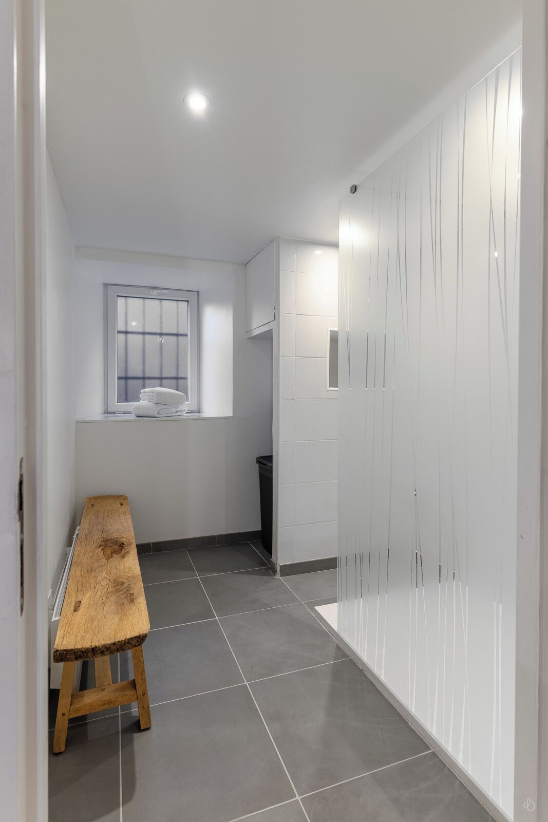 Um corredor com um banco de madeira, uma janela, uma lixeira e um guarda-roupa branco.