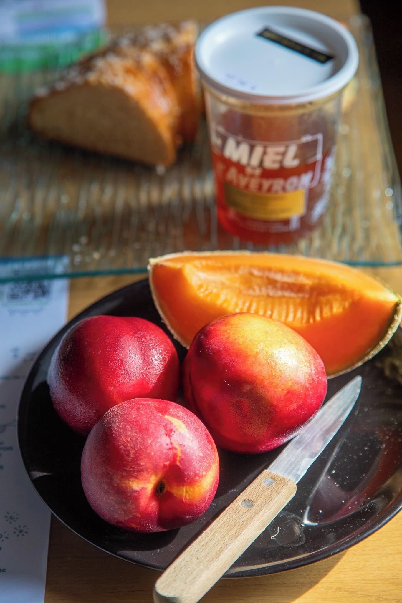 A plate with three peaches and a half-cut cantaloupe, a knife, and a jar of honey on a table.