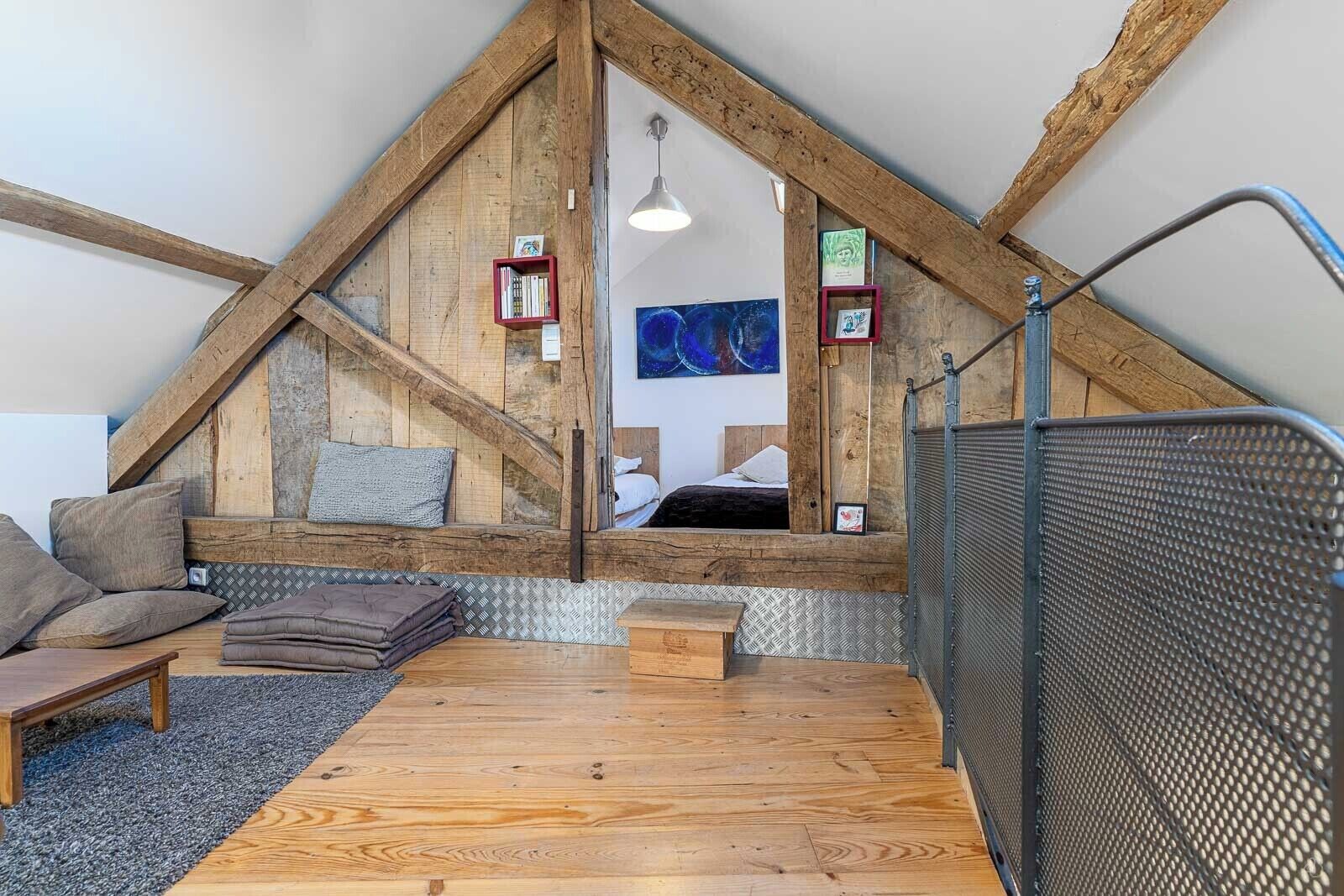 A loft-style bedroom with a sloped ceiling, wooden beams, a bed, a small table, and a seating area with cushions.