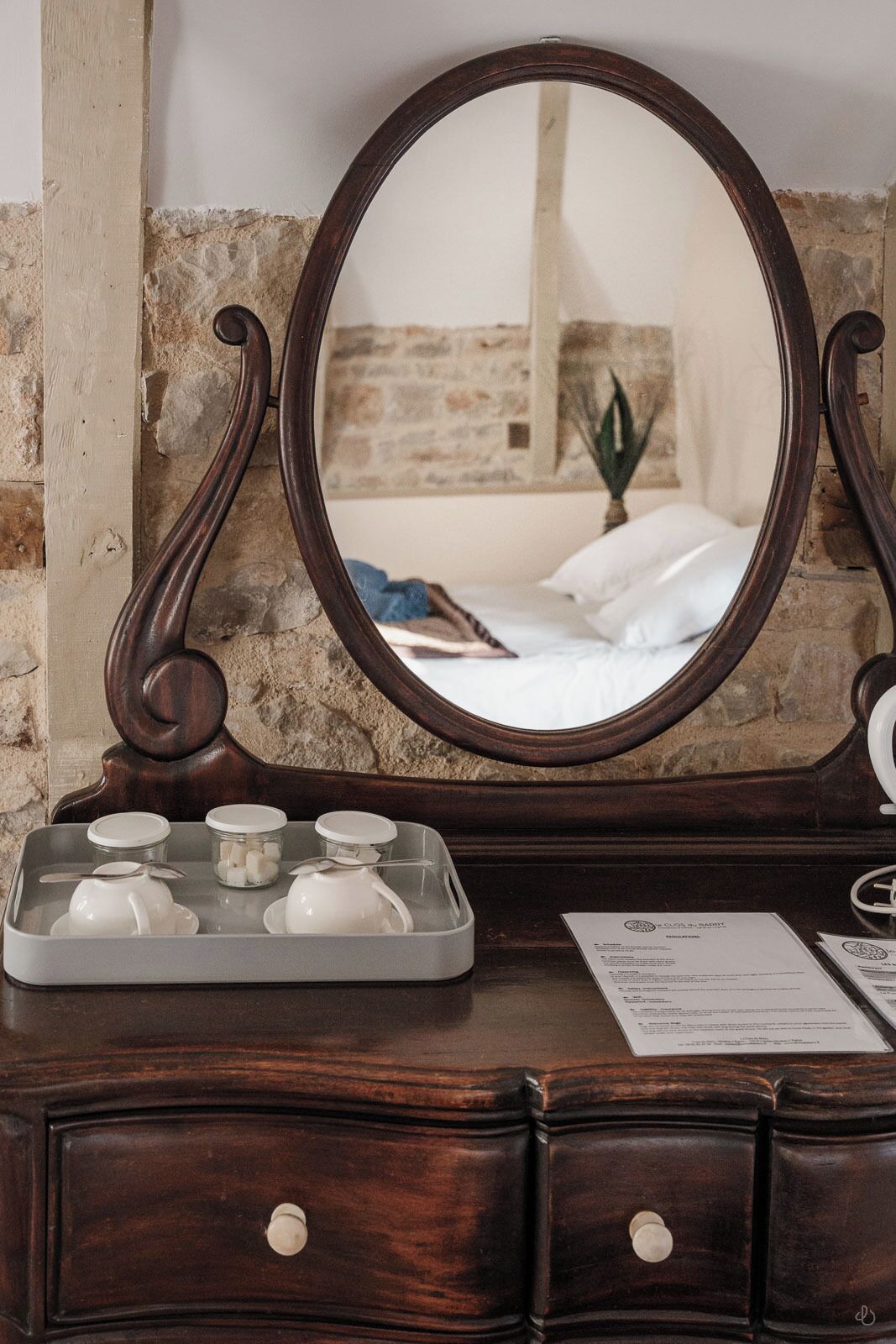 A wooden dresser with a mirror, a tray with cups and sugar cubes, and a bed with white linens in the background.