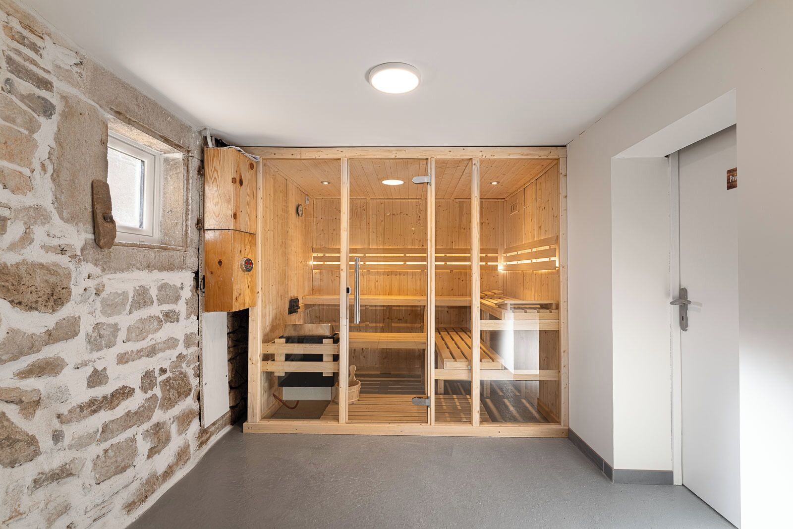 Una sala de sauna con paredes de madera y una puerta de cristal, una pared de piedra contigua y una puerta blanca a la derecha.