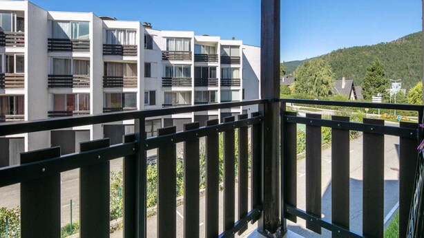 En modern byggnad med flera balkonger, fri utsikt över den omgivande grönskan och en parkeringsplats på förgrunden.