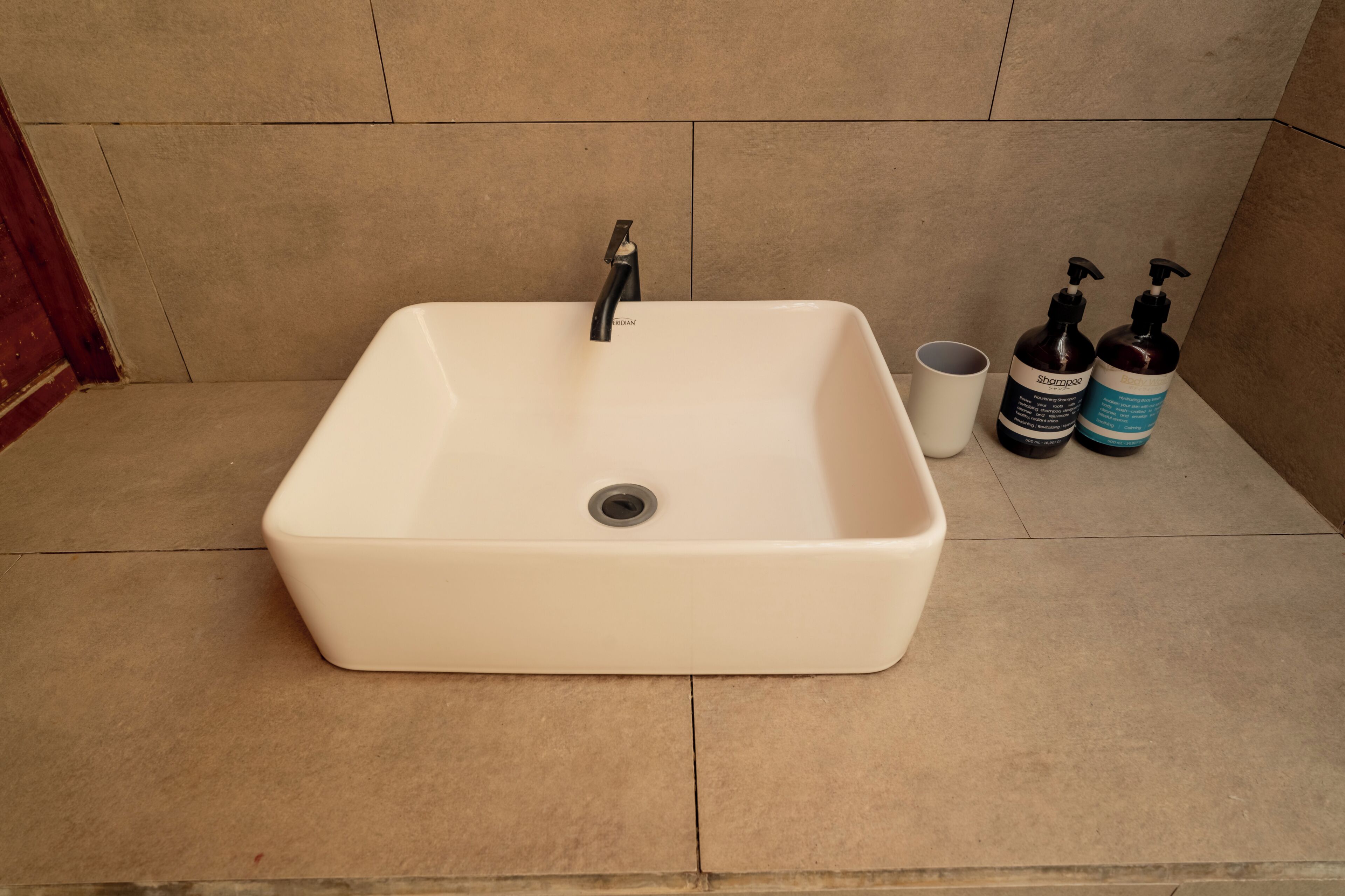 A bathroom with a white sink, a faucet, a cup, and two bottles of shampoo.