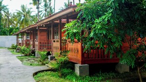 A traditional wooden house with a porch, surrounded by lush greenery and palm trees.