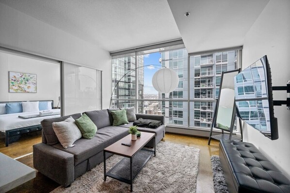 Ein modernes Wohnzimmer mit einem grauen Sofa, einem kleinen Couchtisch und einem großen Fenster mit Blick auf die Stadt.