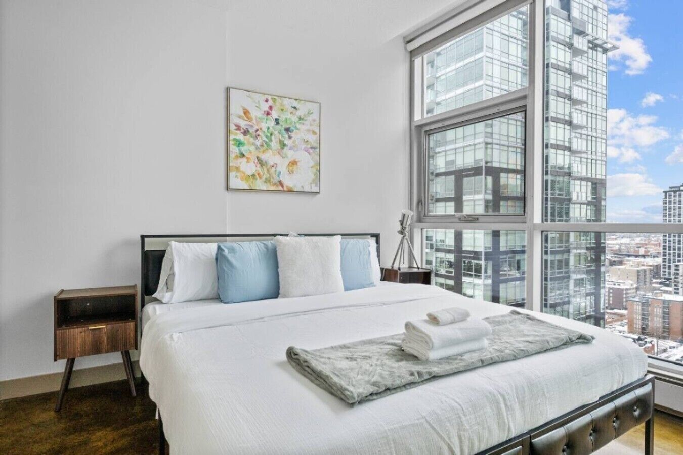 Une chambre d’hôtel moderne avec un grand lit, une table de chevet, une lampe et une vue sur la ville par une grande fenêtre.