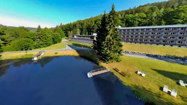 A large building with multiple floors, a body of water in front, and a forested hill in the background.