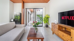 A modern living room with a sofa, a wooden coffee table, a TV, and a sliding glass door leading to a balcony. - Cendana Villa Sanur (Denpasar)