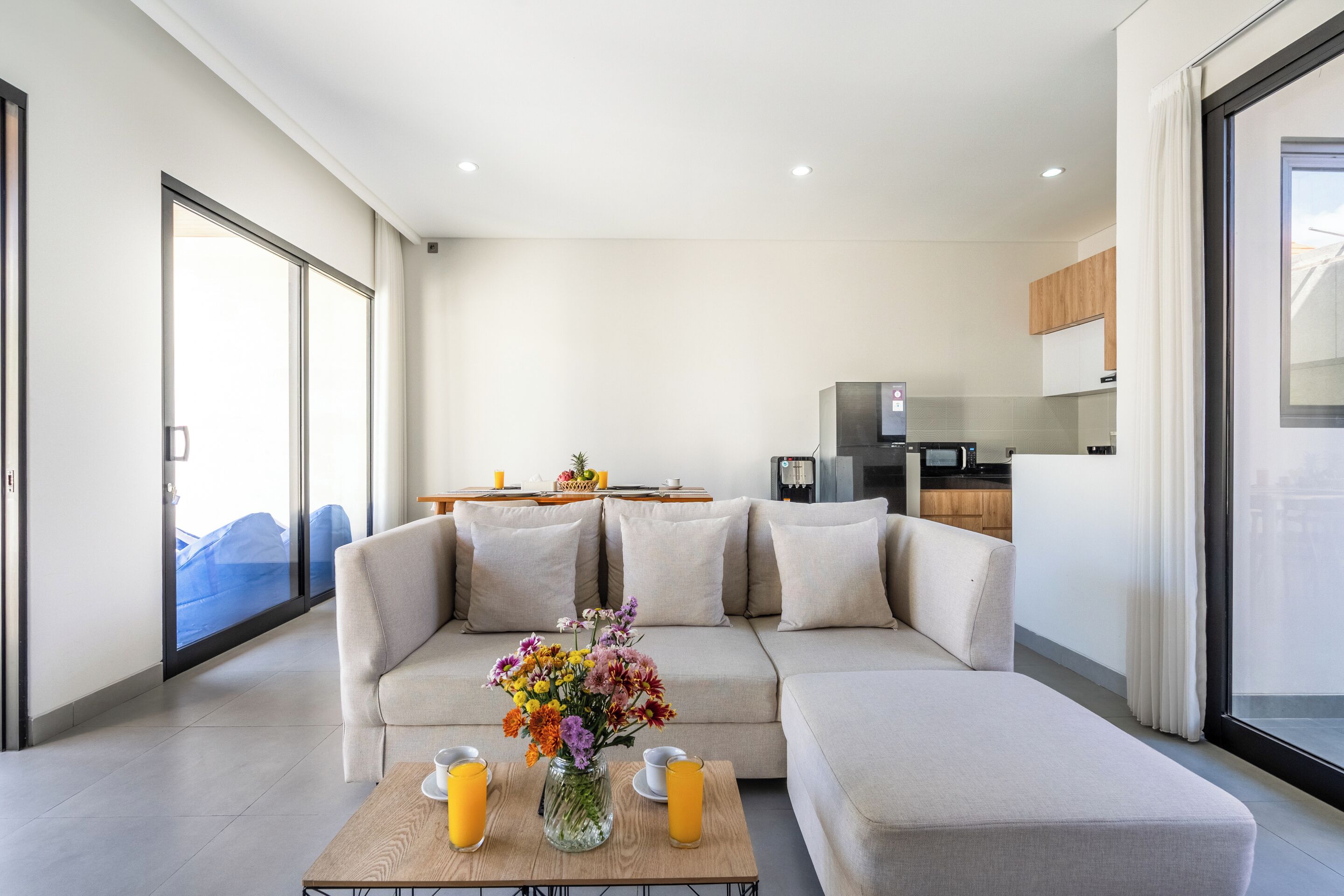 A modern living room with a sofa, a coffee table with drinks and a vase, and a kitchen in the background.