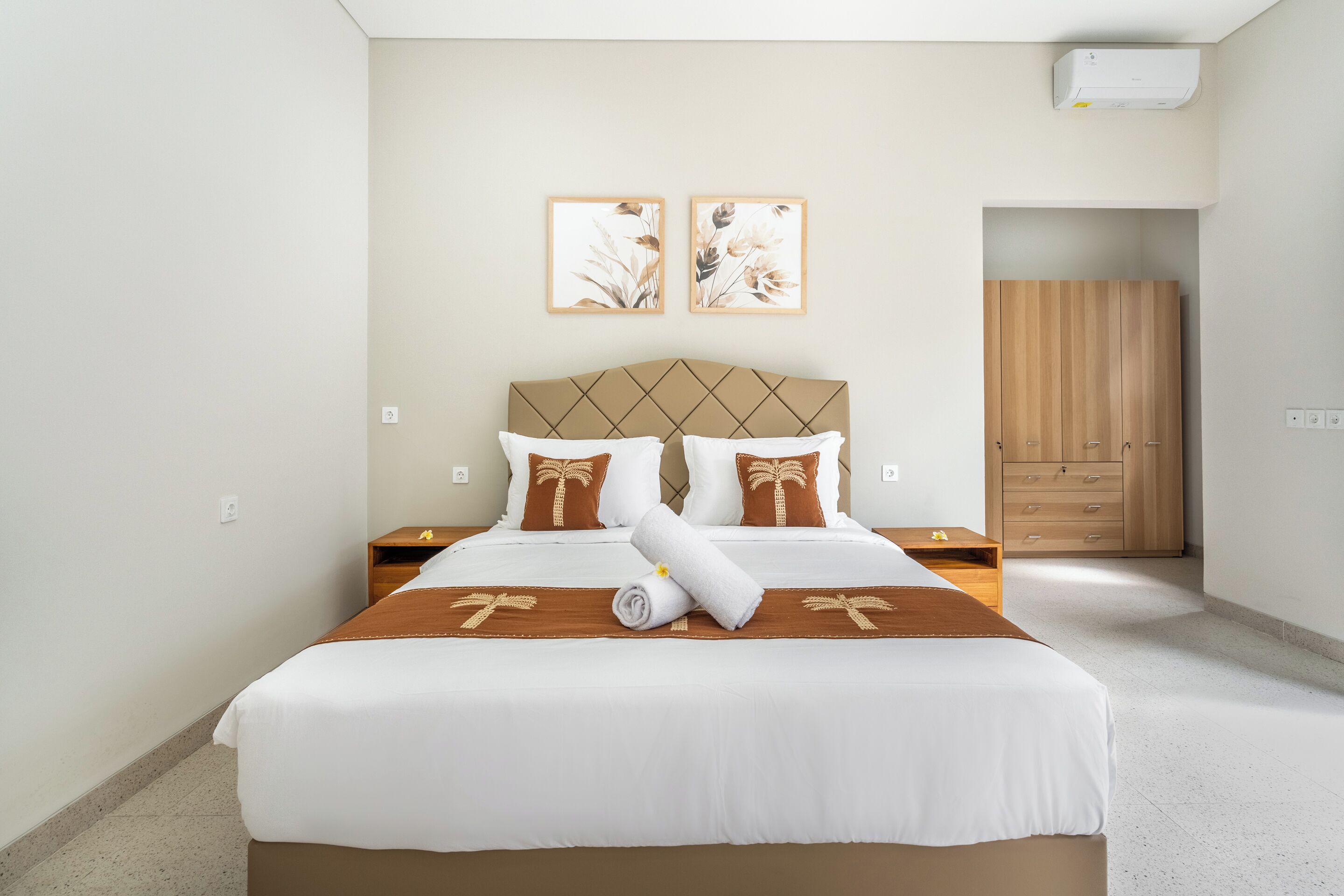 Un lit bien fait, avec une tête de lit marron, du linge de lit blanc et des coussins décoratifs. On aperçoit deux climatiseurs fixés au mur et une armoire en bois à l’arrière-plan.