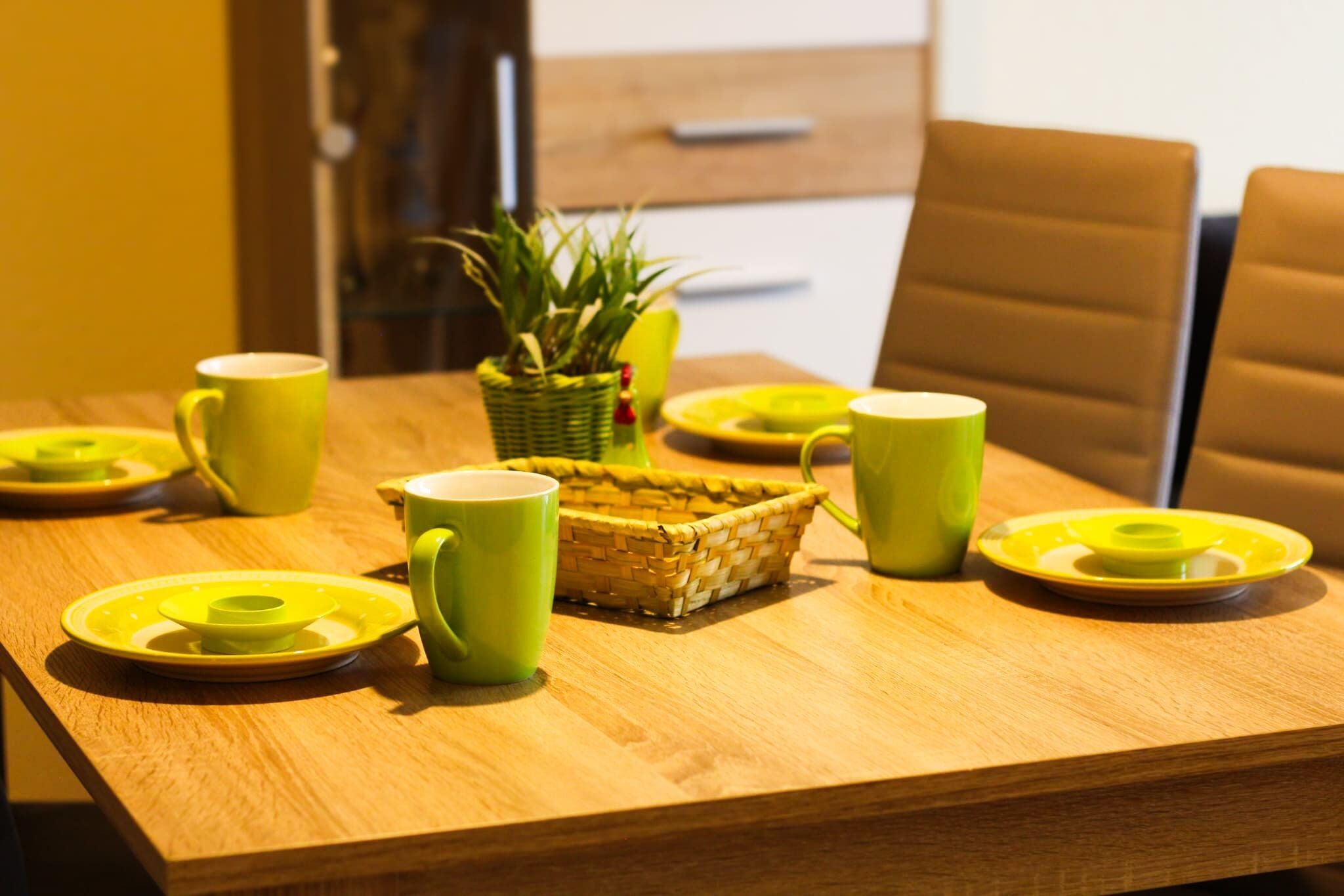 Une table à manger en bois, avec de l’assiette verte et des tasses assorties, un panier en osier et une plante en pot.