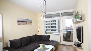 Ett modernt vardagsrum med en hörnsoffa, ett litet bord med en växt, en tv och en balkong med utsikt över byggnader.
