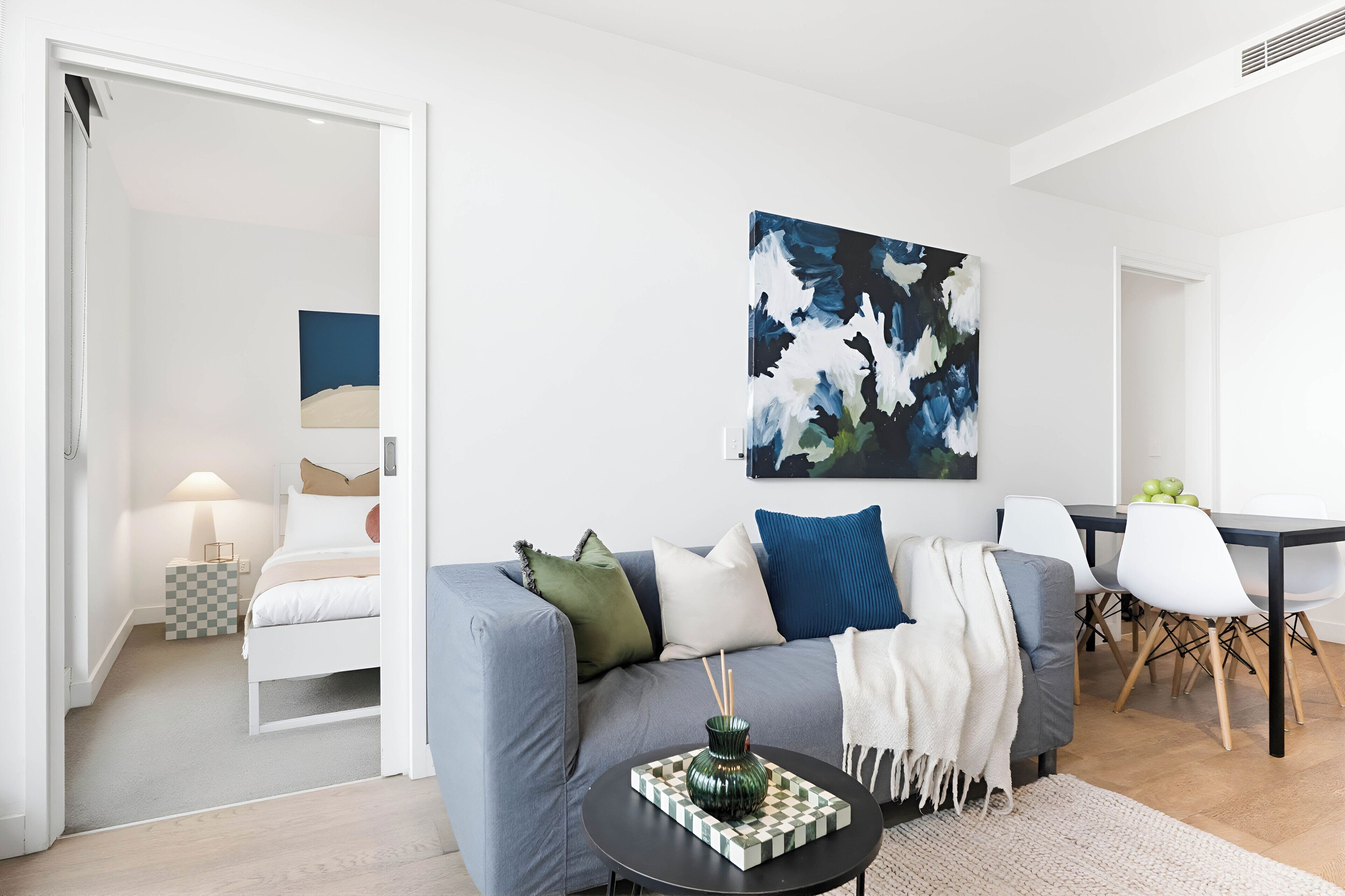 A modern living room with a grey sofa, a dining table with chairs, and a painting on the wall.
