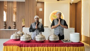 Two individuals standing behind a table with food service items, in a room with wooden paneling and a ceiling fan. - Ambika Hills Resort (Junnar)