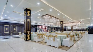 A banquet hall with rows of round tables, white chairs with gold sashes, and a large chandelier. - Nirvana Hotel & Banquet (Batala)