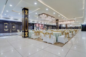 Un salón de banquetes con filas de mesas redondas, sillas blancas con cintas doradas y una gran lámpara de araña.