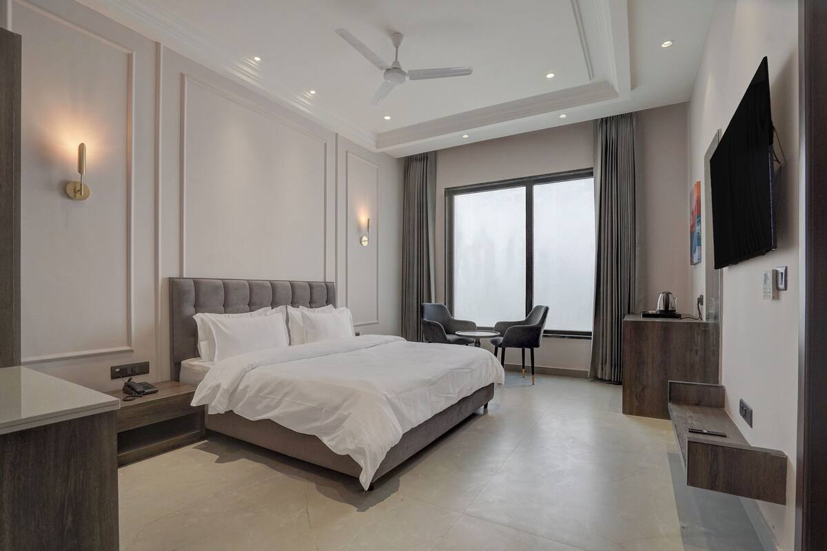 A modern hotel room with a large bed, bedside tables, a desk, a chair, a TV, and a large window with curtains.