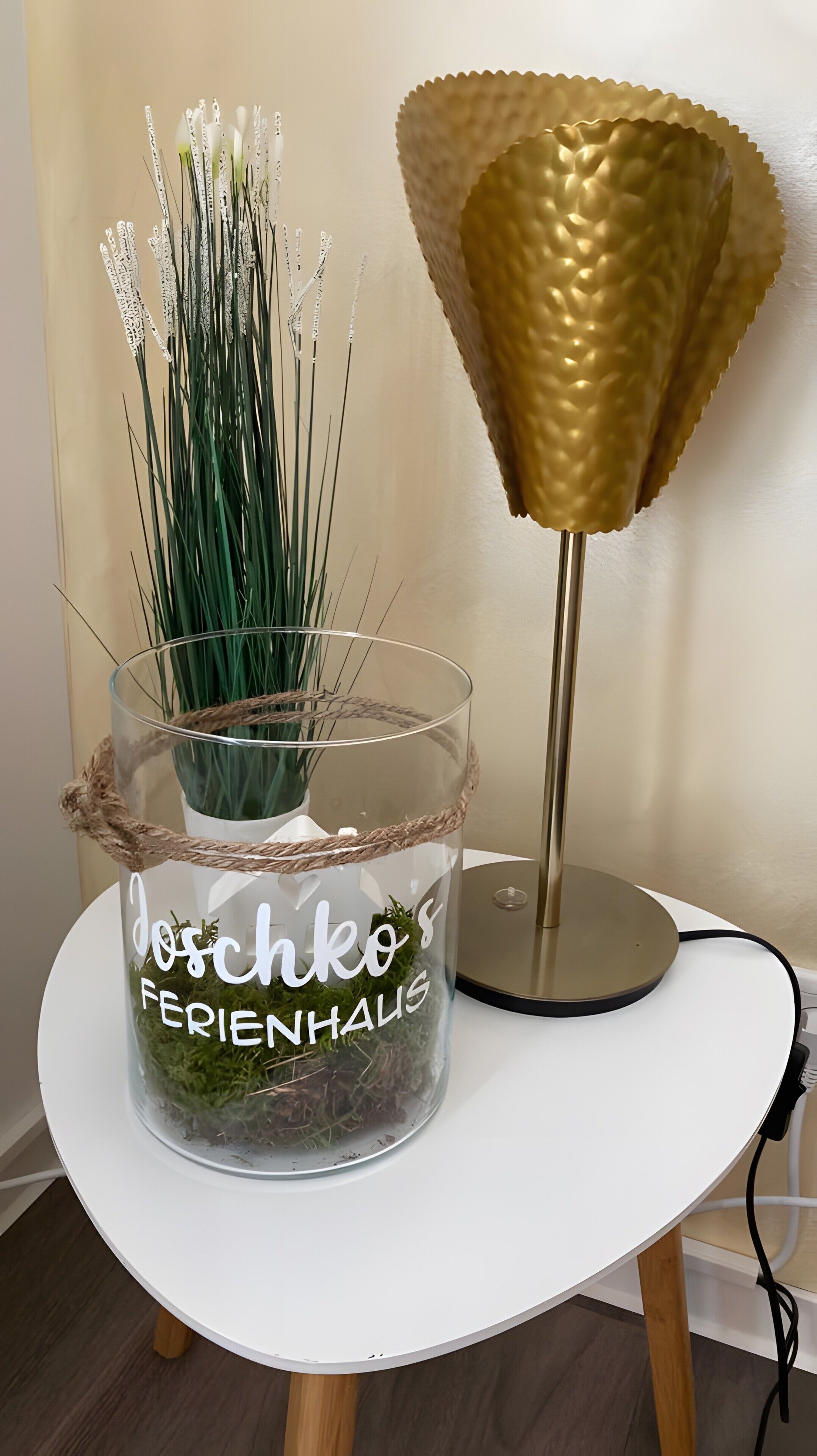 Une table ronde sur laquelle se trouve un vase en verre transparent contenant des plantes vertes et une lampe dorée.