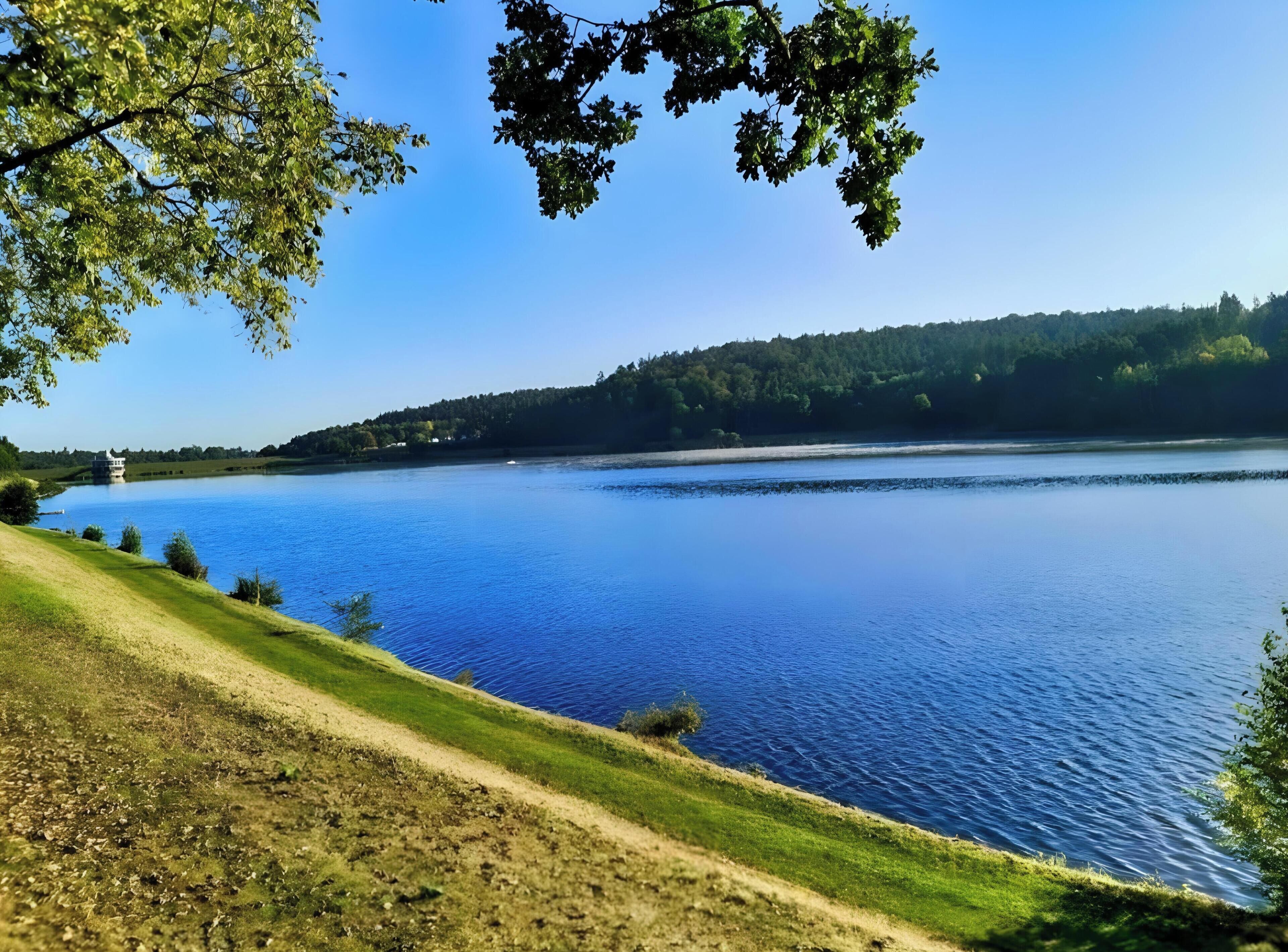 Ein See mit Bootssteg, grüner Uferböschung und einem bewaldeten Hügel im Hintergrund.