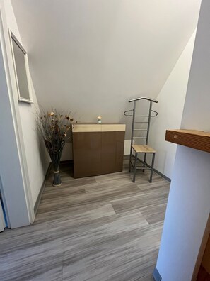 Une pièce d’angle avec un meuble en bois, une chaise en métal, un vase contenant des fleurs séchées et un miroir accroché au mur.