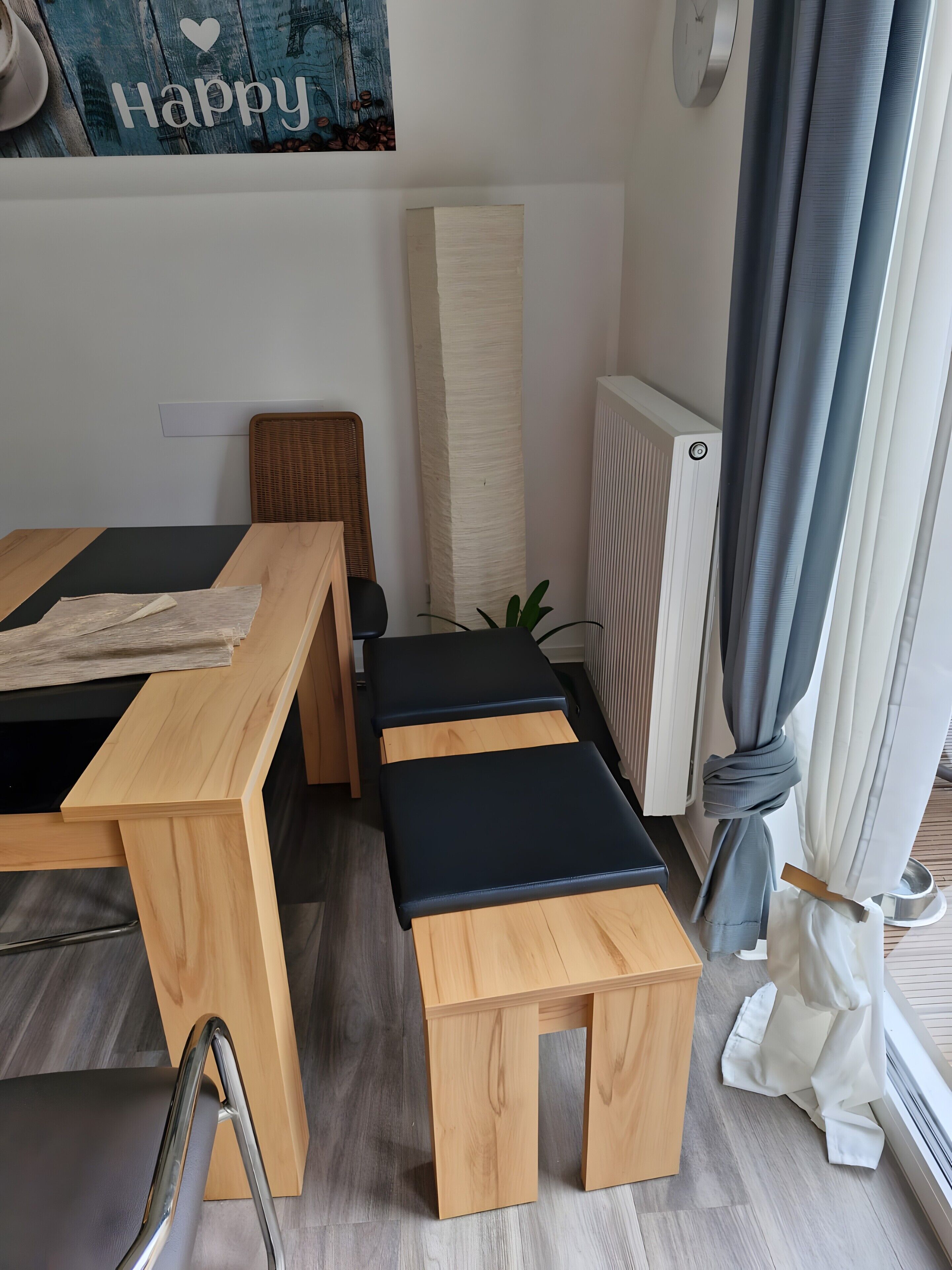 Una habitación con una mesa de madera, dos bancos acolchados negros, una silla de mimbre, un radiador y una ventana con cortinas.