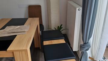 Una habitación con una mesa de madera, dos bancos acolchados negros, una silla de mimbre, un radiador y una ventana con cortinas.