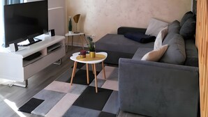 Un salon moderne avec un téléviseur à écran plat, un canapé gris, une petite table ronde et un luminaire suspendu au plafond.