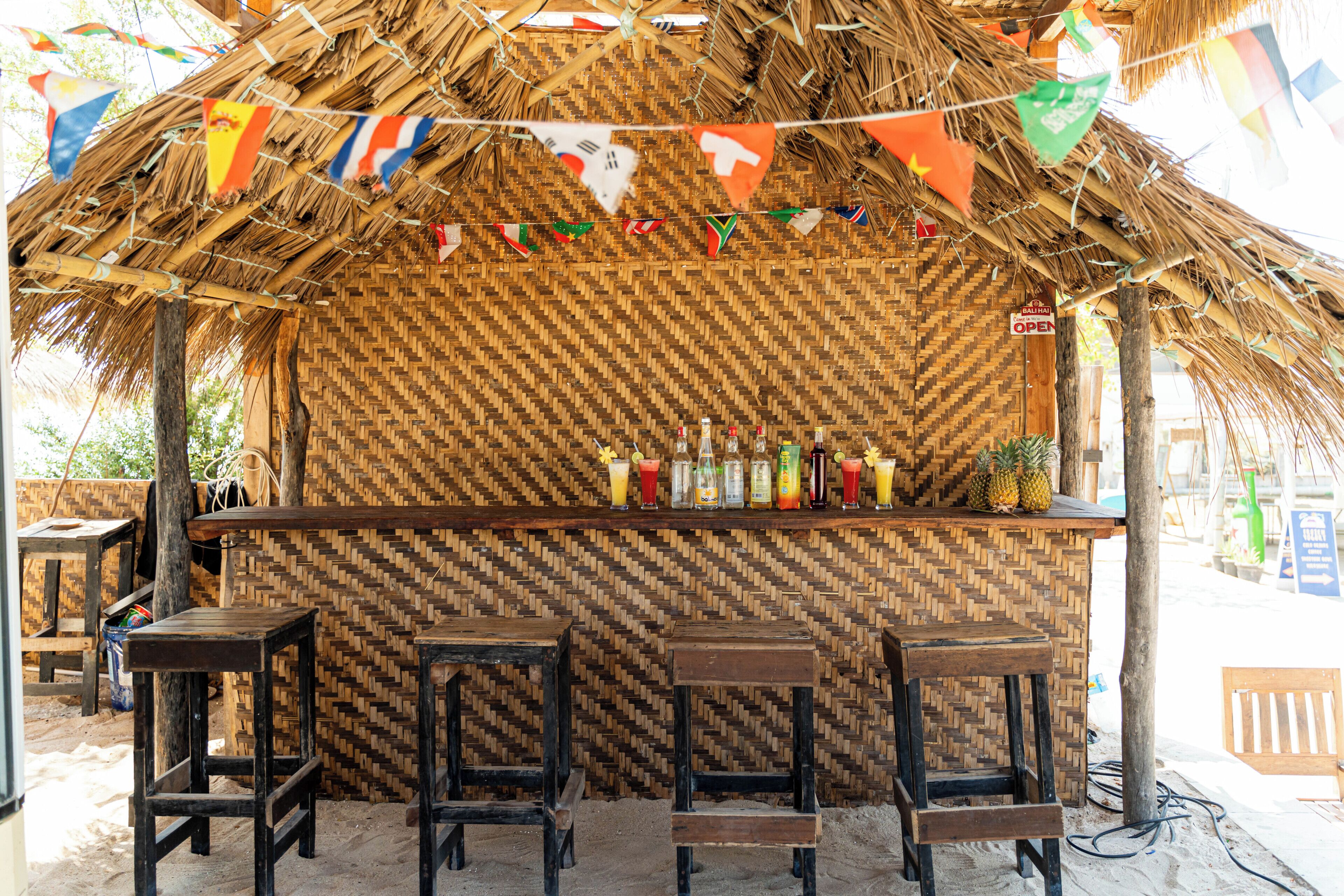 Zona de bar con techo de paja, pared con motivo de espiga, taburetes y una variedad de botellas y vasos sobre la barra.
