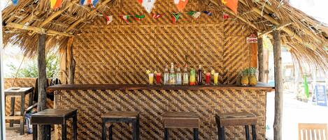 Zona de bar con techo de paja, pared con motivo de espiga, taburetes y una variedad de botellas y vasos sobre la barra.