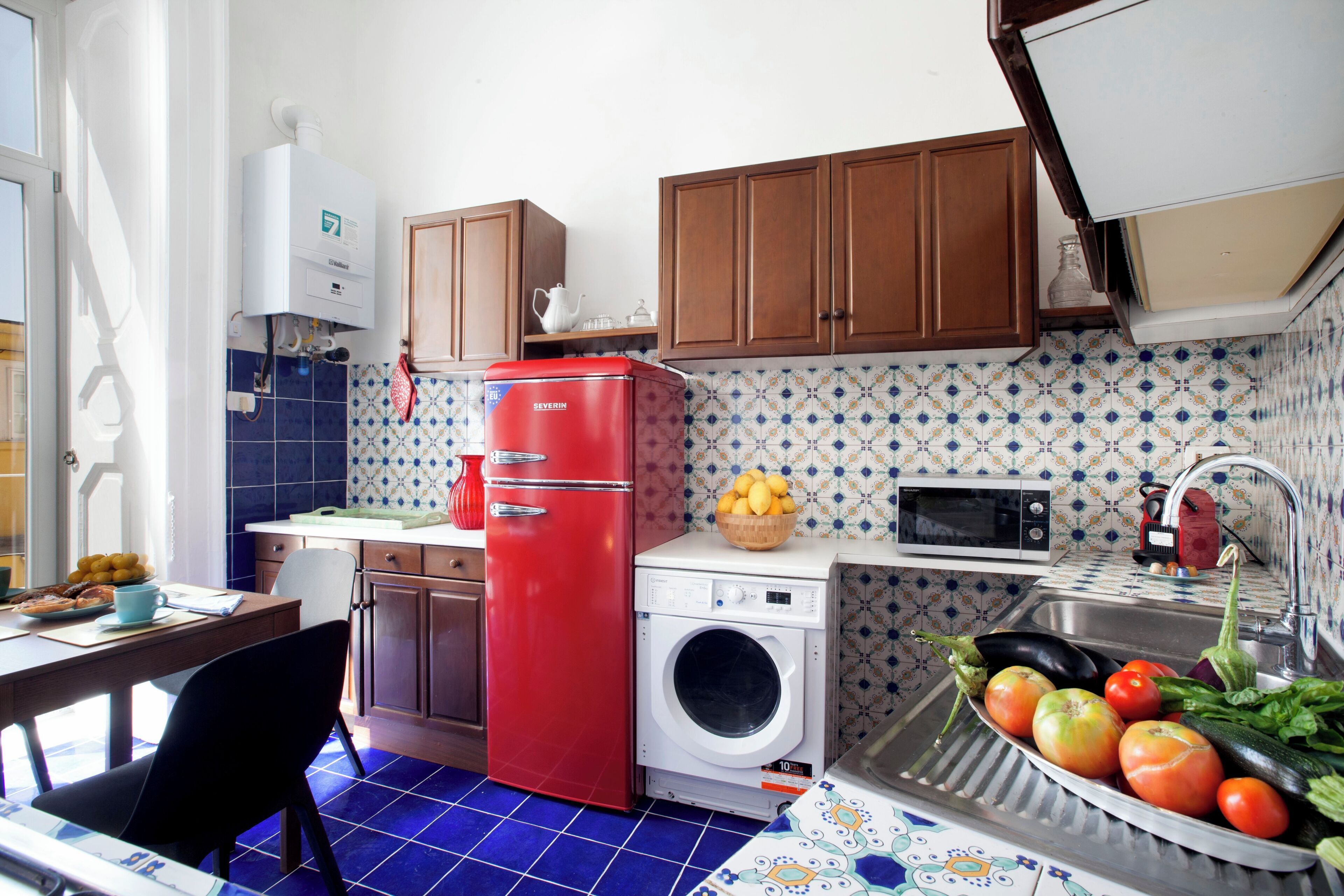 A compact kitchen with a washing machine, a red refrigerator, a microwave, and a sink with fresh vegetables.