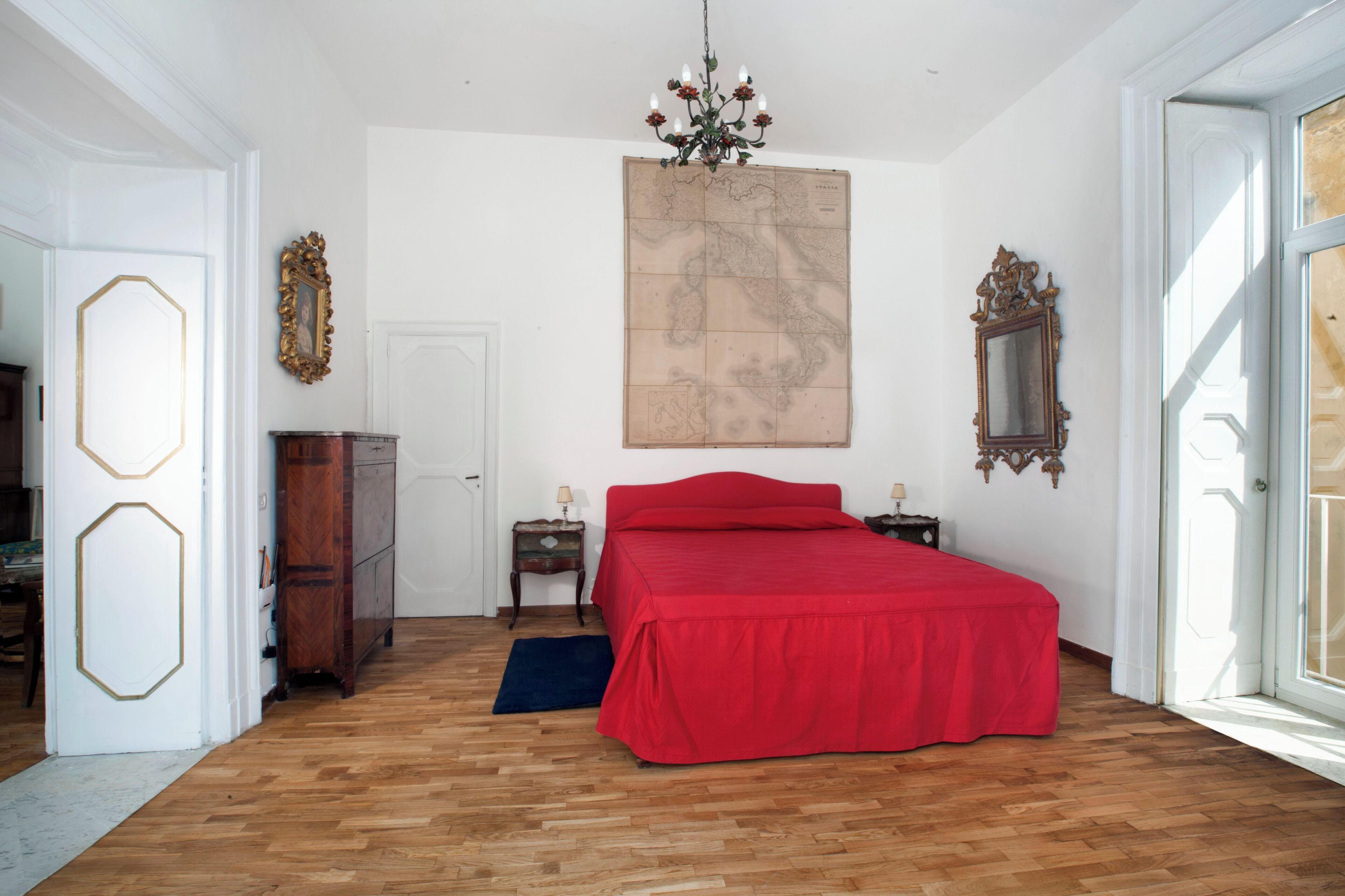 A bedroom with a wooden floor, a red bed, a map on the wall, and a chandelier.
