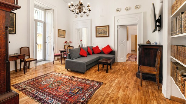 A spacious living room with a grey sofa, red cushions, a patterned rug, wooden flooring, a chandelier, and a bookshelf. - Teresella Suite (Naples)