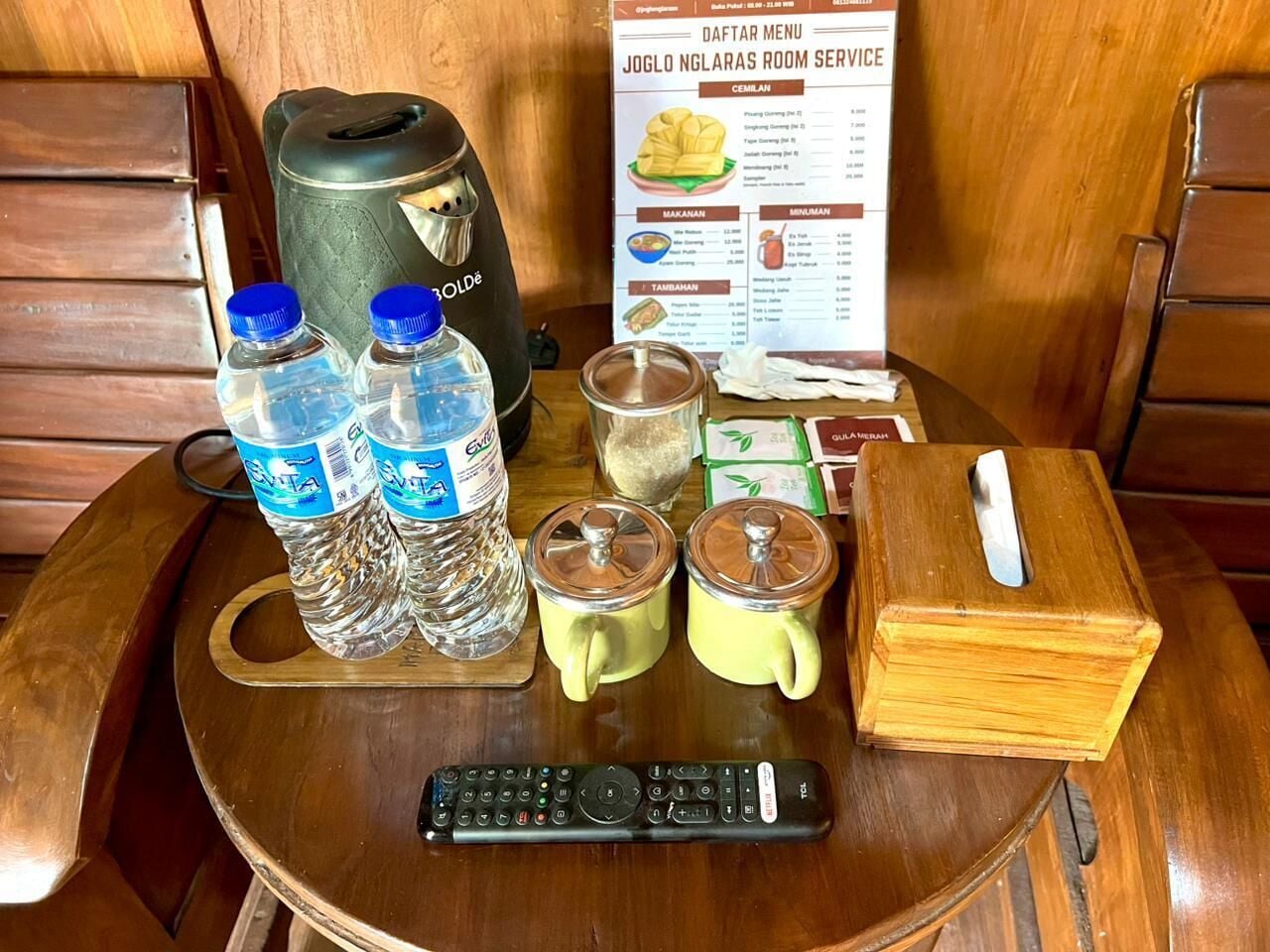 Una mesa de madera con un control remoto, botellas de agua, una tetera, una taza y un menú.