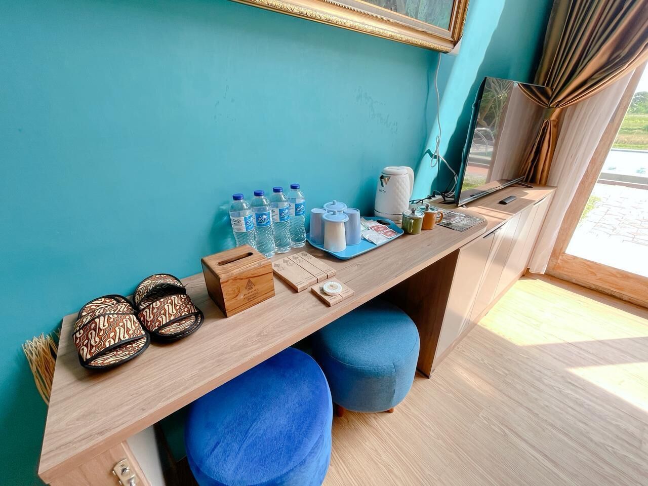 A room with a wooden desk, blue ottomans, a mirror, and various items like slippers, bottles, and a kettle.