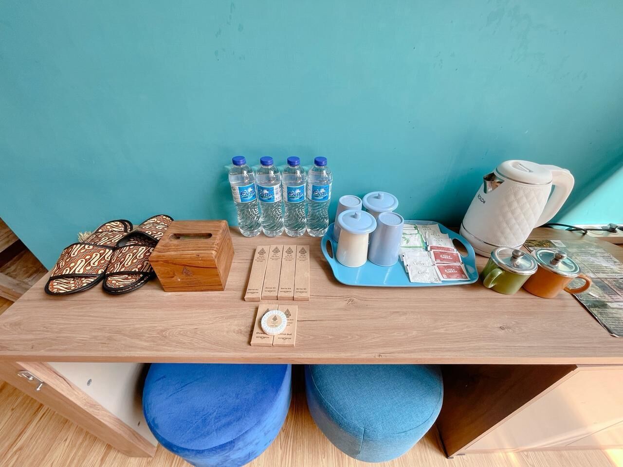 Ein Holztisch mit blauem Kissen, ein Paar gemusterter Hausschuhe, eine Holzkiste, eine Ablage mit Kaffee- und Teebeuteln und mehrere Wasserflaschen.
