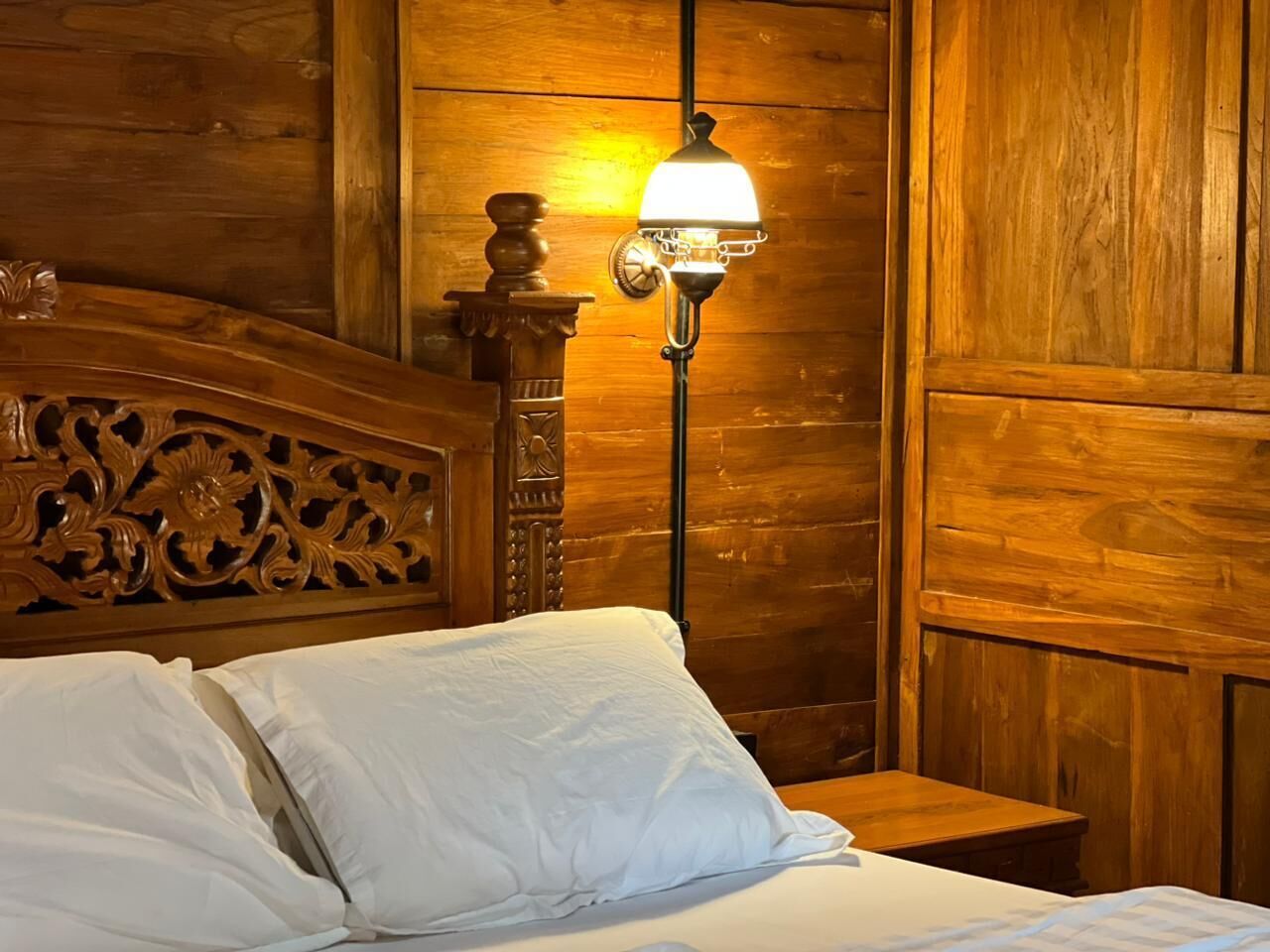 A wooden bed with white linens, a nightstand, and a wall-mounted lamp.