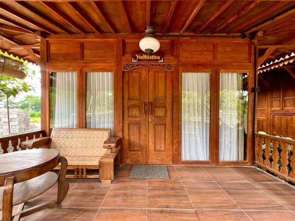 Une véranda en bois avec un banc et une table, une porte en bois surmontée d’une enseigne, et une vue sur la verdure à l’extérieur.