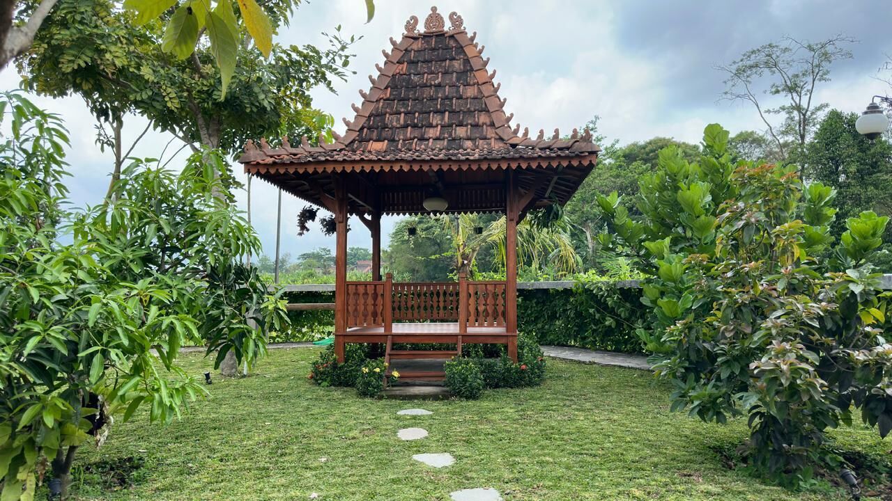 A traditional wooden gazebo with a tiled roof, surrounded by lush greenery and a stone pathway leading to it.